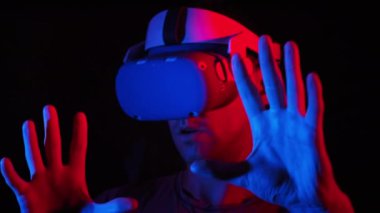 Young man in VR helmet interacts with virtual reality and media content. Male plays a game in VR headset under red-blue light. Use virtual reality futuristic technology. Immersion in metaverse concept