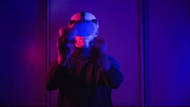 Genç adam sanal gerçeklikte VR kulaklık ve kumanda kullanarak bir oyun oynuyor. Erkek, kırmızı-mavi ışıkta VR kaskındaki 360 medya içeriğiyle etkileşime giriyor. Fütürist teknoloji. Meta-evrene daldırma kavramı