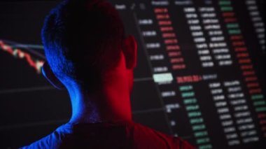 Trader analyze cryptocurrency charts on a big screen in the dark. Rear view of man in red-blue lighting looking at currency charts in stock market. Trading stocks, bitcoins, futures, investment market