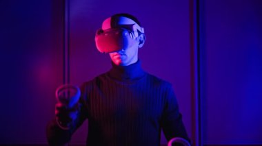 VR kasklı genç adam sanal gerçeklikle kontrolörleri kullanarak etkileşime giriyor. Erkek oyun oynuyor ya da kırmızı-mavi ışıkta VR kulaklıkla 360 medya içeriğini görüyor. Fütürist teknoloji. Meta-evrene daldırma kavramı