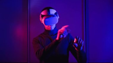 VR kasklı genç adam ellerini kullanarak sanal gerçeklikle etkileşime giriyor. Erkek oyun oyunları, kırmızı-mavi ışık altında VR kulaklıkla 360 medya içeriği. Fütürist teknoloji. Meta-evren kavramına daldırma