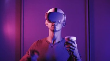 Young man in VR helmet interacts with virtual reality using controllers. Male plays game or see 360 media content in VR headset at red-blue light. Futuristic technology. Concept immersion in metaverse