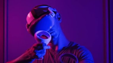 Young man in VR helmet interacts with virtual reality using controllers. Male plays game or see 360 media content in VR headset at red-blue light. Futuristic technology. Concept immersion in metaverse