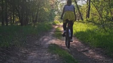 Güneşli bir yaz gününde yeşil orman yolunda bisiklet süren genç bir kadın. Ağır çekim. Bisikletli bir kadın kırsalda yeşil çimlerin çok yakınında patikaya çıkıyor. Sağlıklı bisiklet yaşam tarzı, boş zaman aktivitesi.