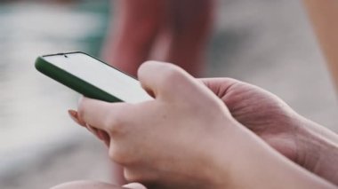 Genç bir kadın sahilde akıllı telefon kullanıyor. Plajda oturan mayo giymiş güzel bir kız elinde bir cep telefonu tutuyor ve sosyal ağları izliyor. Yaşam tarzı. Deniz kıyısındaki eğlence aktiviteleri