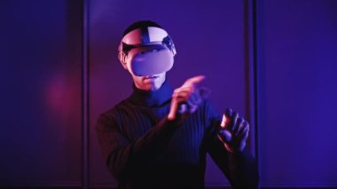 Young man in a VR helmet interacts with virtual reality and media content. Concept of male plays game in VR headset at red-blue light. Use virtual reality futuristic technology. Immersion in metaverse