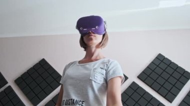 A young woman in a VR helmet plays a game at home. Emotional Female using virtual reality futuristic headset and controllers play in a 3D game. Spending modern leisure. Immersion in the metaverse.