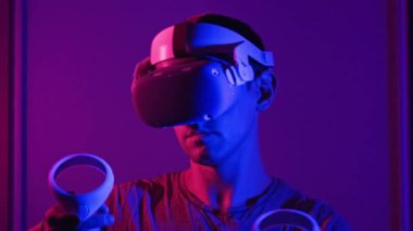 VR kasklı genç adam sanal gerçeklikle kontrolörleri kullanarak etkileşime giriyor. Erkek oyun oynuyor ya da kırmızı-mavi ışıkta VR kulaklıkla 360 medya içeriğini görüyor. Fütürist teknoloji. Meta-evrene daldırma kavramı
