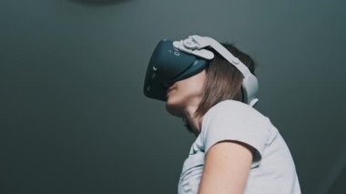 Young woman in a virtual reality headset plays a game at home. Emotional female using VR futuristic helmet and controllers play a game in 360. Spending modern leisure. Immersion in the metaverse. 4K