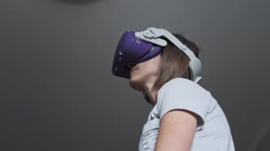 Sanal gerçeklik kulaklığı takmış genç bir kadın evde oyun oynuyor. VR fütüristik kask kullanan duygusal kadın ve kontrolörler 360 'da bir oyun oynuyorlar. Modern eğlenceyi harcamak. Meta-evrene dalma. 4K