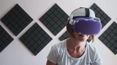 Sanal gerçeklik kulaklığı takmış genç bir kadın evde oyun oynuyor. VR fütüristik kask kullanan duygusal kadın ve kontrolörler 360 'da bir oyun oynuyorlar. Modern eğlenceyi harcamak. Meta-evrene dalma. 4K