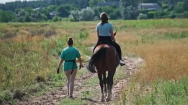 Back view of young woman riding a horse on a path near green nature, horseback riding, slow motion. Pretty girl learning horseback riding on a summer day in countryside. The horsewoman is on a saddle.