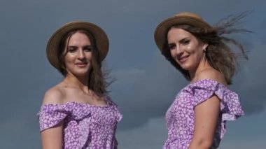 Portrait of two young twin girls in identical summer dresses and straw hats looking at the camera in nature. Smiling twin sisters in the countryside stand on the field against the sky. Slow motion