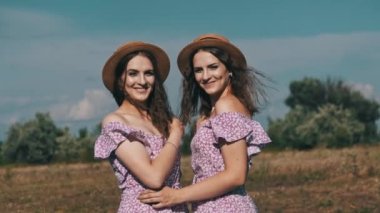 Portrait of two smiling young twin women in summer dresses and straw hats looking at the camera in nature. Attractive identical sisters in the countryside stand on field against the sky. Slow motion