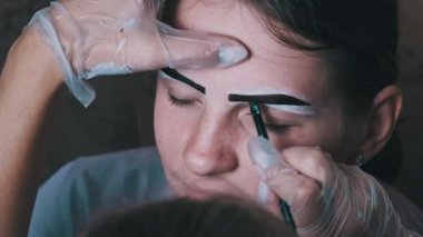 Eyebrow tinting procedure with henna in a beauty salon. The master brow corrects the shape of the eyebrows with a brush. A professional cosmetologist makes make-up. Face of a young girl close-up. 4K