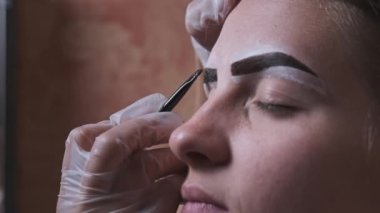 Building the shape of the eyebrows. Beauty salon. Eyebrow master dyes them for a young woman. Eyebrow correction and tint procedure. Girl sits with natural brown henna on brows. Close-up face. 4K