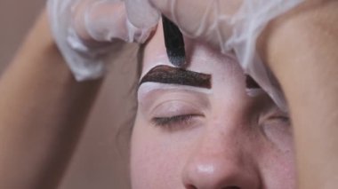 Building the shape of the eyebrows. Beauty salon. Eyebrow master dyes them for a young woman. Eyebrow correction and tint procedure. Girl sits with natural brown henna on brows. Close-up face. 4K