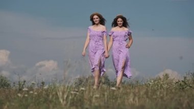 Yeşil tarlada el ele tutuşan yaz elbiseli ve hasır şapkalı güzel ikizler. Yaşam tarzı. Aynı mor elbiseli iki çekici kadın yavaş çekimde çayırdan geçiyor.