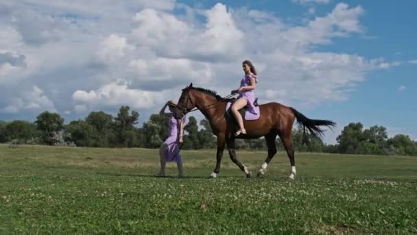 スローモーションで緑のフィールドで馬に乗って歩く若い双子の姉妹 夏のドレスやわらの帽子で若い双子の女性は 1つの乗馬 他の近くの馬をリードしています 乗馬ライフスタイル — ストック動画