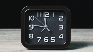 Ahşap masa ve siyah arkaplan üzerindeki işaretli saat zamanı gösteriyor. İkinci el, kare şeklindeki analog saatin yuvarlak kahverengi kadranı boyunca hareket eder. Zaman yönetimi, günlük rutin, iş, motivasyon