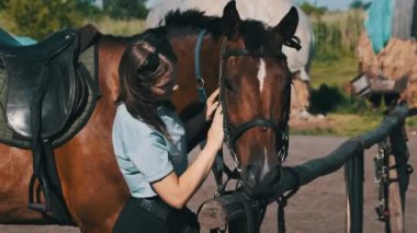 Happy woman stroking a horse on a farm in nature at summer day. Slow Motion. Beautiful girl pets brown horse near the stable. Friendship between human and animal. Mutual understanding, love for horses