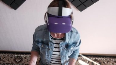 A young woman in a VR helmet plays a game at home. Emotional Female using virtual reality futuristic headset and controllers play in a 3D game. Spending modern leisure. Immersion in the metaverse.