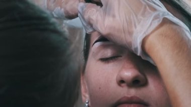 Eyebrow tinting procedure with henna in a beauty salon. The master brow corrects the shape of the eyebrows with a brush. A professional cosmetologist makes make-up. Face of a young girl close-up. 4K