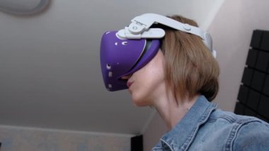 VR kasklı genç bir kadın evde oyun oynuyor. Sanal gerçeklik fütüristik kulaklık kullanan duygusal kadın ve kontrolörler üç boyutlu bir oyun oynuyorlar. Modern eğlenceyi harcamak. Meta-evrene daldırma.