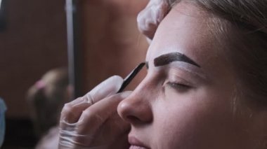 Building the shape of the eyebrows. Beauty salon. Eyebrow master dyes them for a young woman. Eyebrow correction and tint procedure. Girl sits with natural brown henna on brows. Close-up face. 4K