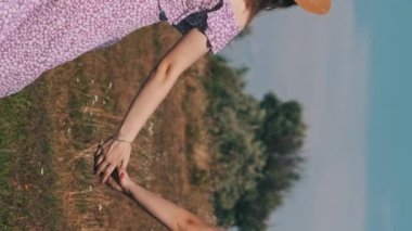 Vertical, Back view of young twins sisters in summer dresses and straw hats holding hands walking by a green field. Attractive two women in identical dresses together going by meadow in slow motion