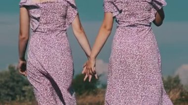 Back view of young twins sisters in summer dresses and straw hats holding hands walking by a green field. Attractive two women in identical purple dresses together going by a meadow in slow motion