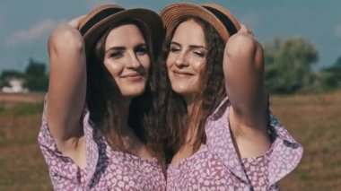 Aynı yaz elbiseleri ve hasır şapkalı iki genç ikiz kız doğada kameraya bakıyor. Kırsalda gökyüzüne karşı duran gülümseyen ikiz kız kardeşlerin portresi. Yavaş çekim