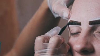 Building the shape of the eyebrows. Beauty salon. Eyebrow master dyes them for a young woman. Eyebrow correction and tint procedure. Girl sits with natural brown henna on brows. Close-up face. 4K