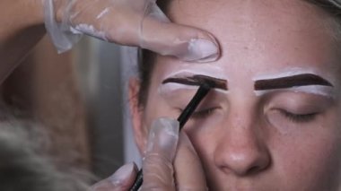 Building the shape of the eyebrows. Beauty salon. Eyebrow master dyes them for a young woman. Eyebrow correction and tint procedure. Girl sits with natural brown henna on brows. Close-up face. 4K