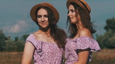 Portrait of two young twin girls in identical summer dresses and straw hats looking at the camera in nature. Smiling twin sisters in the countryside stand on the field against the sky. Slow motion