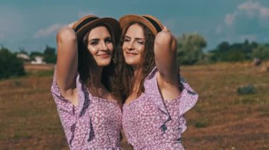 Portrait of two smiling young twin women in summer dresses and straw hats looking at the camera in nature. Attractive identical sisters in the countryside stand on field against the sky. Slow motion