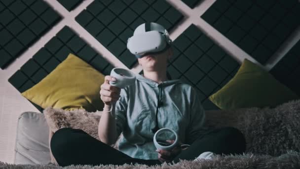 穿着虚拟现实头盔的年轻女子坐在沙发上 手里拿着操纵杆回家 在室内使用Vr头盔玩游戏 穿着Vr模拟器的女孩享受3D网络空间 Vr眼镜 — 图库视频影像