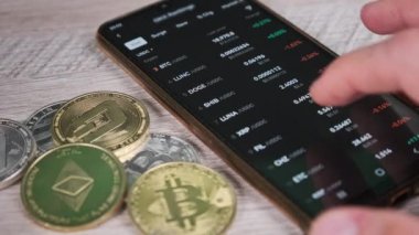 Tüccar, bitcoin ve kripto paraların olduğu bir masada akıllı telefon ekranındaki kripto para fiyatlarını analiz ediyor. Bir sürü şifreli bilgi kaynağı. İş adamı dijital piyasayı inceliyor. Mal varlığının yüzde büyümesi veya düşüşü