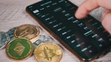 Mobil takas uygulamasındaki akıllı telefon ekranındaki şifreli para birimlerinin listesi. Kripto pazarının analizi. Mans el şifreli paralar ve bitcoinlerle dolu bir masada telefon kullanıyor. Birçok şifreli varlık