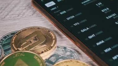 Mobil takas uygulamasındaki akıllı telefon ekranındaki şifreli para birimlerinin listesi. Kripto pazarının analizi. Mans el şifreli paralar ve bitcoinlerle dolu bir masada telefon kullanıyor. Birçok şifreli varlık