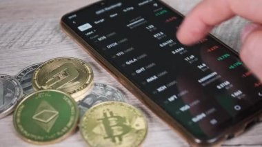 Tüccar, bitcoin ve kripto paraların olduğu bir masada akıllı telefon ekranındaki kripto para fiyatlarını analiz ediyor. Bir sürü şifreli bilgi kaynağı. İş adamı dijital piyasayı inceliyor. Mal varlığının yüzde büyümesi veya düşüşü