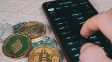Mobil takas uygulamasındaki akıllı telefon ekranındaki şifreli para birimlerinin listesi. Kripto pazarının analizi. Mans el şifreli paralar ve bitcoinlerle dolu bir masada telefon kullanıyor. Birçok şifreli varlık