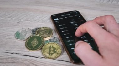 Tüccar, bitcoin ve kripto paraların olduğu bir masada akıllı telefon ekranındaki kripto para fiyatlarını analiz ediyor. Bir sürü şifreli bilgi kaynağı. İş adamı dijital piyasayı inceliyor. Mal varlığının yüzde büyümesi veya düşüşü