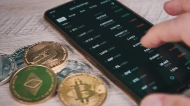 Mobil takas uygulamasındaki akıllı telefon ekranındaki şifreli para birimlerinin listesi. Kripto pazarının analizi. Mans el şifreli paralar ve bitcoinlerle dolu bir masada telefon kullanıyor. Birçok şifreli varlık