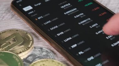 Mobil takas uygulamasındaki akıllı telefon ekranındaki şifreli para birimlerinin listesi. Kripto pazarının analizi. Mans el şifreli paralar ve bitcoinlerle dolu bir masada telefon kullanıyor. Birçok şifreli varlık
