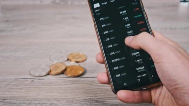 Mobil takas uygulamasındaki akıllı telefon ekranındaki şifreli para birimlerinin listesi. Kripto pazarının analizi. Mans el şifreli paralar ve bitcoinlerle dolu bir masada telefon kullanıyor. Birçok şifreli varlık