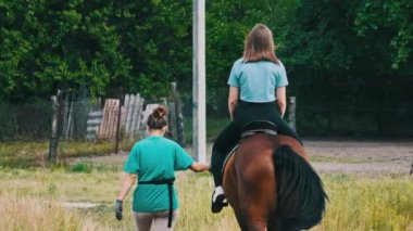 Kırsalda at süren, ata binen, ağır çekimde at süren genç bir kadının arka görüntüsü. Öğretmen kız binicinin yanında yürür. Ata binme dersi. Atlı kadın eyerin üstünde..