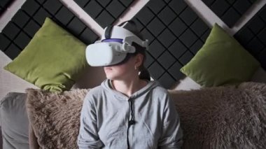 Sanal gerçeklik kaskı takan genç bir kadın kanepede oturuyor ve evde elinde joysticklerle oturuyor. Kapalı alanda video kask kullanarak oyun oynamak. VR simülatörü takan kız 3 boyutlu siber uzayın tadını çıkarıyor. VR gözlükleri