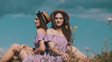 Two twin girls sit together on a field in identical dresses and straw hats. Slow motion. Portrait of happy young twins sisters looking in the camera on a summer meadow in the countryside. Lifestyle