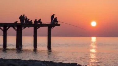 Silhouettes of Fishermens with Fishing Rods at Sea Sunset Sitting on the Pier. TimeLapse. Orange sky. Fisherman catches a fish off the pier. Lifestyle. Time lapse.